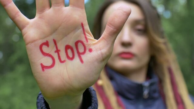 Modern serious girl with long dreadlocks is showing hands with written slogan 'Stop' on green tree background. Responsibility, climate change, protests concepts