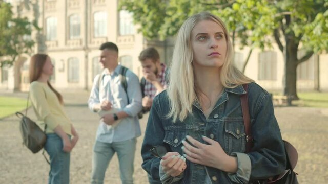 Beautiful shy blond girl smiling at camera as people laughing at her at the background. Confident intelligent Caucasian female student ignoring mockery from groupmates. Intelligence, confidence.