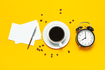 Cup of black coffee, coffee beans, black alarm clock, pens, white paper cards on yellow background Flat lay top view copy space. Creative Concept time to work, female desktop, coffee background