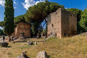 Fototapeta na wymiar Perspective view of the two brick tombs on the Via Appia Antica, Rome. The road was the most important of the Roman Empire, remains of other tombs, rich vegetation, maritime pines, cypresses.