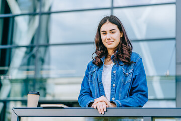 Portrait of young female millennial student in jeans jacket outdoor with modern urban building on background. Hispanic girl freelancer relaxing on coffee break.