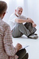 Sad grey senior man sitting on the floor during group psychotherapy for men