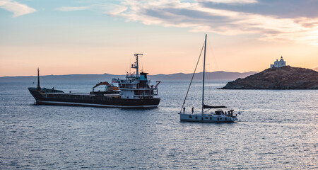 Obraz premium Sailboat and a ship on calm sea at sunset in the Aegean sea, Greece. Lighthouse on a cape at Kea island.