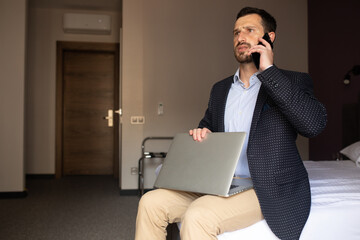 Staying in touch with office. Handsome young man in suit working on laptop and smiling while sitting on the bed in hotel room.