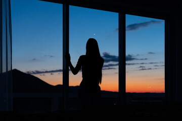 Silhouette of a back of a girl standing near the window and looking through the window dreaming or thinking