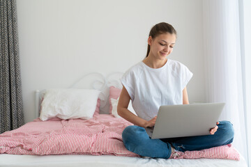 Beautiful girl using laptop and smiling while sitting on bed and studying at home