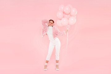 Full length portrait of young girl wearing fur coat, ear warmers and glasses, holding birthday balloons for party, isolated on pink background