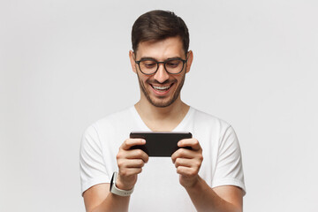 Young man holding phone in horizontal way as if watching funny video, isolated on gray background