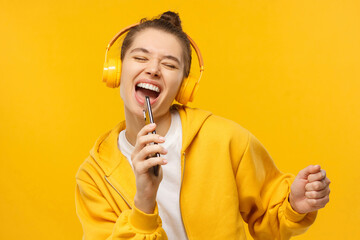 Young girl in yellow hoodie and wireless headphones, singing with closed eyes, holding tphone as...