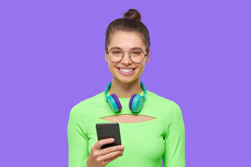 Smiling girl, wearing green neon top, round glasses and wireless headphones around neck, holding phone in hands, isolated on purple background