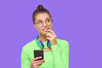 Young dreamful girl in glasses, neon green top and with headphones around neck, looking away and dreaming, isolated on purple background