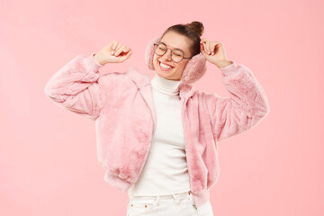 Excited girl wearing fur coat, earmuffs and round glasses, dancing to sound of music, isolated on pink background