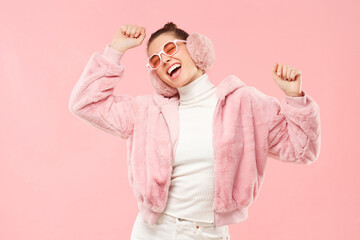 Young excited girl wearing warm furry coat, earmuffs and colored eyeglasses, moving arms to sounds of music, isolated on pink background - obrazy, fototapety, plakaty