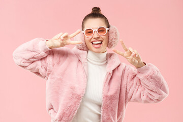 Happy smiling young girl in cozy winter coat, earmuffs and colored glasses, dancing and showing victory sign, isolated on pink background