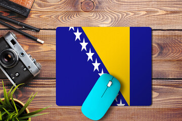 Bosnia Herzegovina flag on wooden background with blue wireless mouse on a mouse pad, top view....