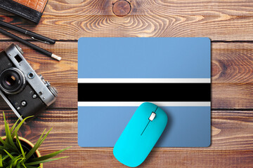 Botswana flag on wooden background with blue wireless mouse on a mouse pad, top view. Digital media concept.