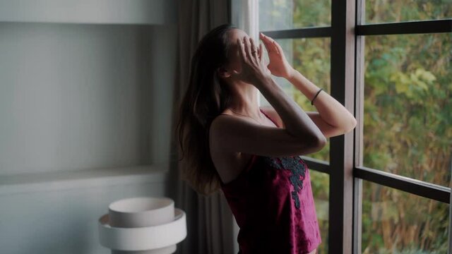 Woman in pajamas wakes up, enjoys view from window and ties ponytail in her hair