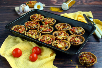 Baked tartlets with vegetables on a baking sheet and fresh tomatoes on a dark wooden table. Closeup