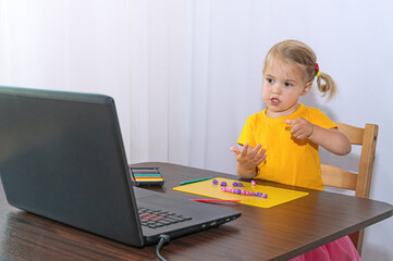 Naklejka premium An interested little girl looks at her laptop and sculpts out of plasticine. Interested girl in an online lesson next to her laptop.