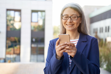 Checking email. Portrait of cheerful mature business woman in classic wear using smartphone and smiling while standing against office building outdoors