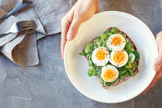 Hard boiled egg on avocado toast with green leaves, healthy breakfast or lunsh, top view, man's hands holding a plate, grey background
