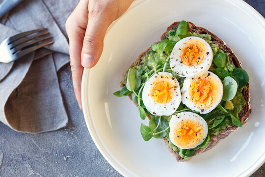 Hard boiled egg on avocado toast with green leaves, healthy breakfast or lunsh, top view, man's hand holding a plate