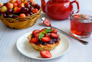 Appetizing cupcake with strawberries, red teapot, cup and basket with ripe cherries on a light wooden table