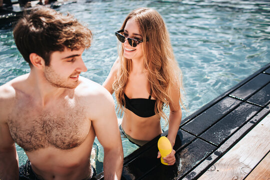 Young hot couple resting at swimpool. Sexy amazing beautiful man and woman together in water posing for photo camera. Girl n sunglasses looks at guy and smiles. Holding suncream yellow bottle in hand.
