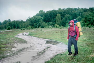 Guy tourist hiker walks along the road with a backpack in a red jacket in the rain