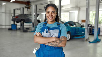 Top service is my motive. Portrait of young african american woman, professional female mechanic in uniform smiling at camera, standing in auto repair shop. Car service, repair, maintenance concept