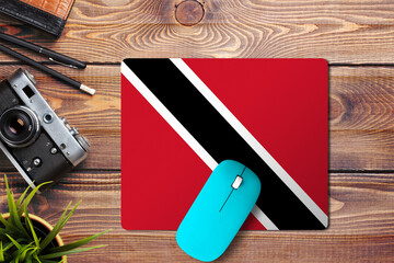 Trinidad And Tobago flag on wooden background with blue wireless mouse on a mouse pad, top view. Digital media concept. © sezerozger