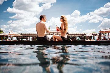 Young hot couple resting at swimpool. A view of man and woman look at each other and drink...