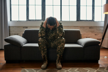 Constant pain. Full length shot of middle aged military man holding his head in pain and depression sitting on the couch. Soldier suffering from psychological trauma. PTSD concept