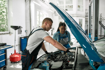 Take control of a car. Caucasian man, professional male mechanic repairing car engine, tighten, screw with spanner while his female colleague holding torch under car hood at service station