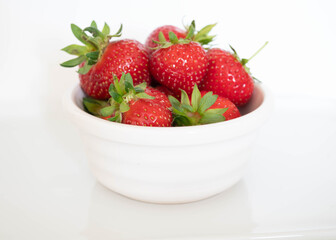 A white bowl of vivid red juicy strawberries with green stalks.  Fresh fruit in a bowl against a white background with copy space