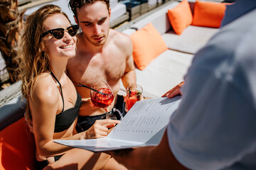 Young hot couple resting at swimpool. Cheerful man and woman choosing food from restaurant menu and look at waiter. Girl smiling. Drinking red cocktails and enjoy rest or relax at spa resort place.
