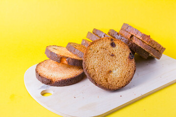 several pieces of skuar with raisins on a wooden cutting board, a tower of round baked loaf of rusks on a yellow background