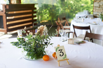 Composition of field herbs and cereals on the table in the restaurant in a transparent vase.White tablecloth and two tangerines. Stillife.