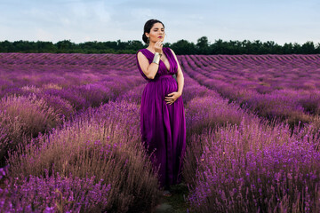Beautiful pregnant woman dressed in a purple dress in a lavender field smells a lavender branch. Pregnancy and motherhood concept.