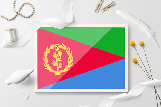 Eritrea flag in wooden frame on white creative background. White theme, feather, daisy, button, ribbon objects.