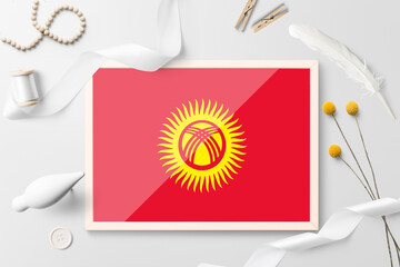 Kyrgyzstan flag in wooden frame on white creative background. White theme, feather, daisy, button, ribbon objects.