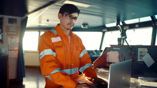 Filipino deck Officer on bridge of vessel or ship wearing coverall during navigaton watch at sea . He is using laptop, electronic paperwork at sea, concept of reporting