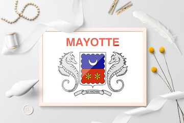 Mayotte flag in wooden frame on white creative background. White theme, feather, daisy, button, ribbon objects.