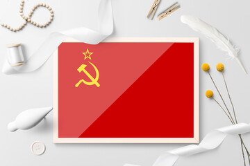 Soviet Union flag in wooden frame on white creative background. White theme, feather, daisy, button, ribbon objects.