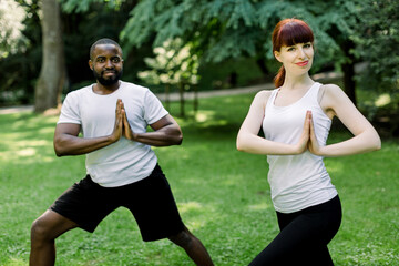 Young multiethnic couple, African man and Caucasian woman, doing yoga in nature outdoors, standing with hands together in prayer position. Healthy lifestyle, practice yoga, meditation concept