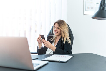 Blonde business woman sitting in office, looking at phone and smiling.