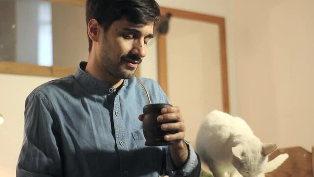 Handsome young man drinking argentine mate and caressing a white cat