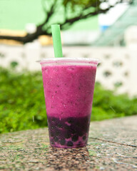 Bright magenta-colored boba drink with green straw sitting on granite ledge with green foliage and white wall in the background. Cinatown in Singapore.
