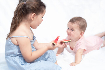 Children eat red watermelon.  The girl treats the baby.  Sisters look at each other