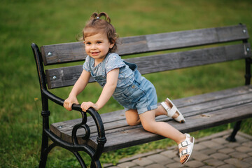 Cute little girl in denim climbs on the bench in the park. Happy smiled kid on the bench